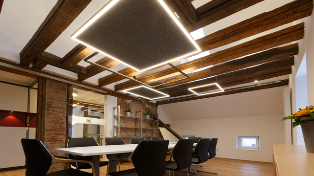 Warm meeting room with a rectangular meeting table with wooden beams on the ceiling and suspended luminaires in various square frame shapes, some fitted with felt acoustic absorbers. | © Kiefer Raimund