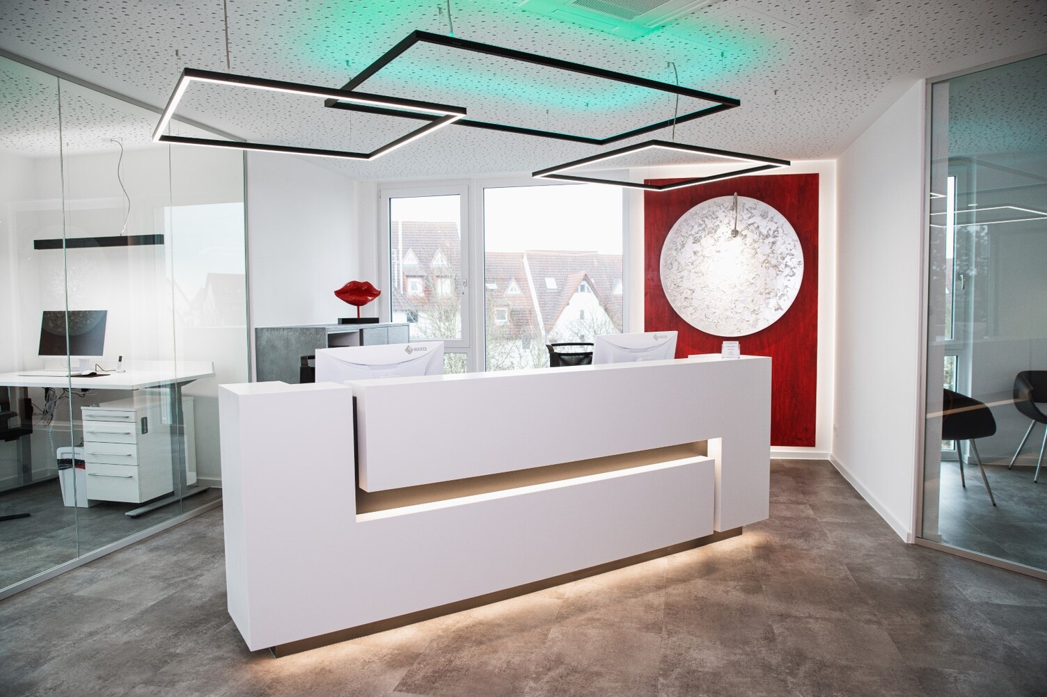 White reception desk at Dr. Hensel's dental practice with windows and a red wall in the background; square pendant luminaires are suspended above the reception desk, two of which shine directly in a white tone and a rectangular one indirectly colours the white ceiling in RGB colours.