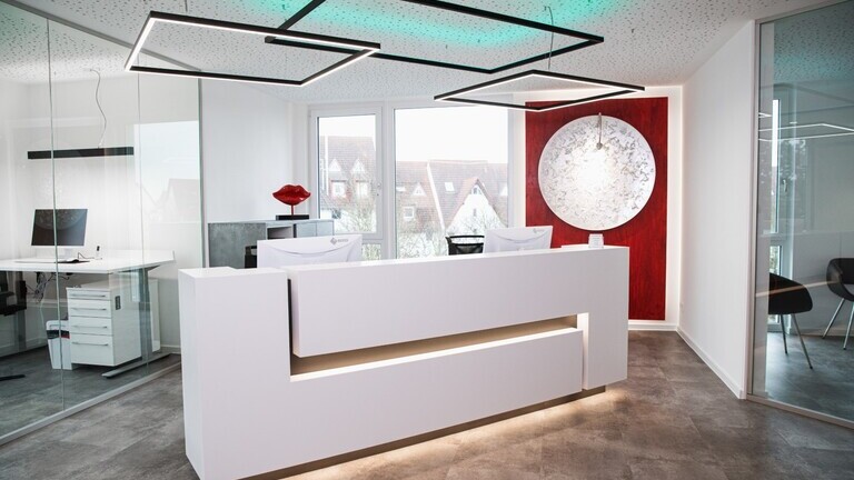 White reception desk at Dr. Hensel's dental practice with windows and a red wall in the background; square pendant luminaires are suspended above the reception desk, two of which shine directly in a white tone and a rectangular one indirectly colours the white ceiling in RGB colours. | © Studio Troncone