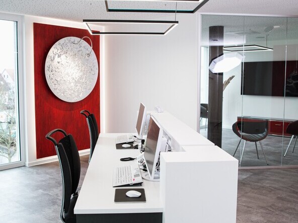 White reception desk at Dr. Hensel's dental practice with windows and a red wall in the background; square pendant luminaires are suspended above the reception desk, two of which shine directly in a white tone and a rectangular one indirectly colours the white ceiling in RGB colours. | © Studio Troncone