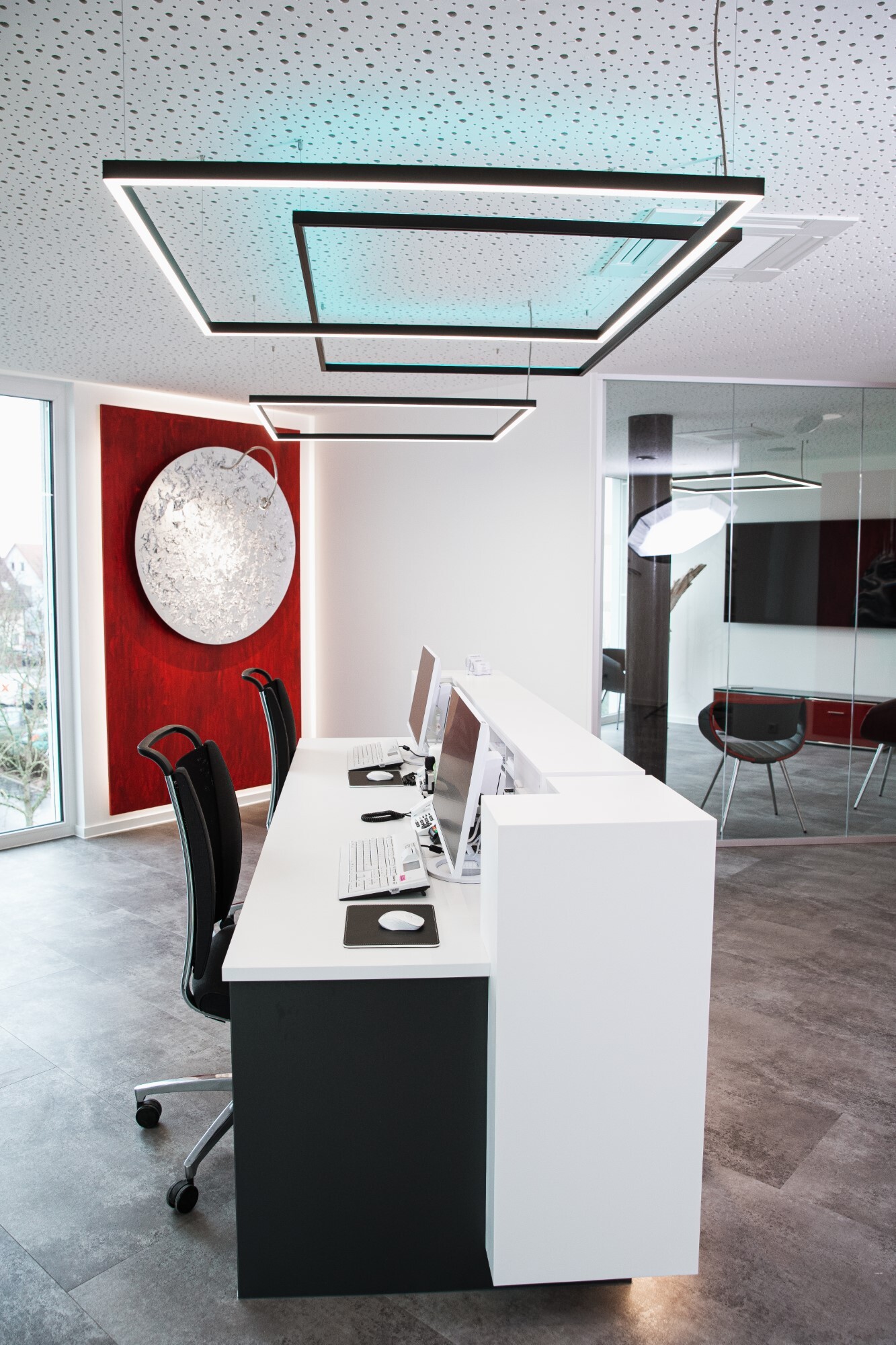White reception desk at Dr. Hensel's dental practice with windows and a red wall in the background; square pendant luminaires are suspended above the reception desk, two of which shine directly in a white tone and a rectangular one indirectly colours the white ceiling in RGB colours.