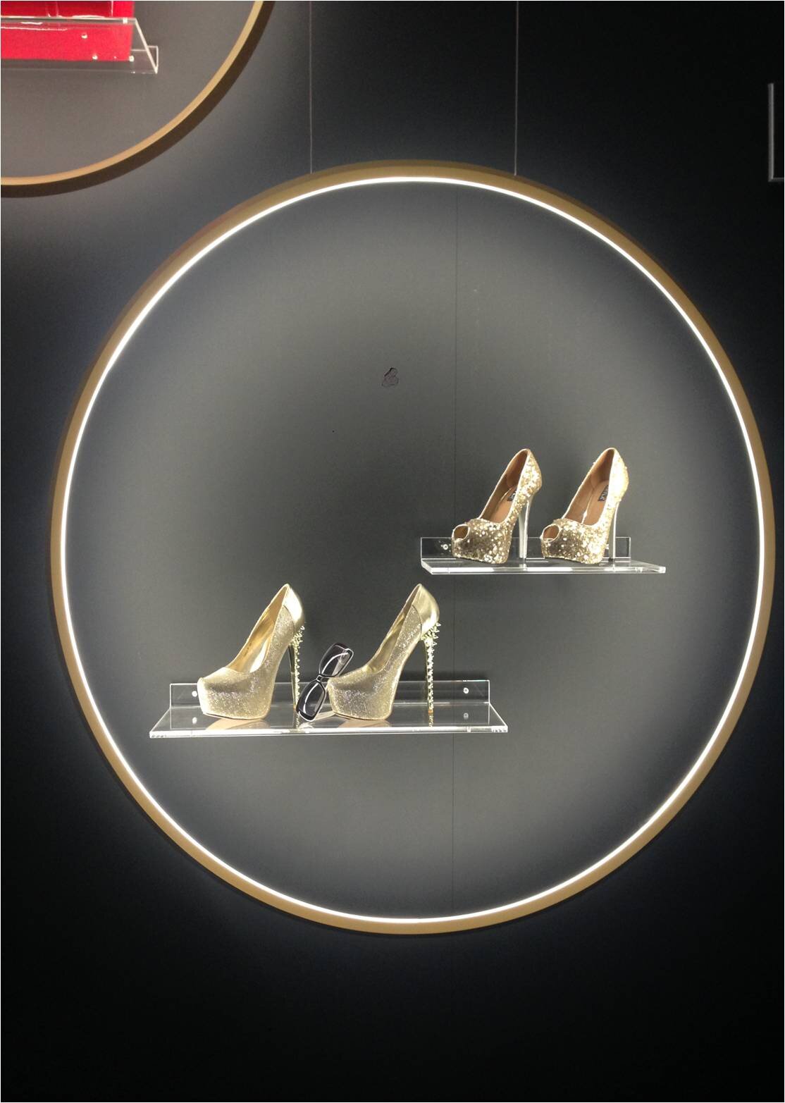 Ringleuchte GIRATA VERTICAL setzt Schuhe in Szene | © SATTLER Innenkreis der Ringleuchte GIRATA als Präsentationsfläche für goldene Schuhe & schwarze Sonnenbrille auf Regalen vor einer schwarzen Wand. | © SATTLER