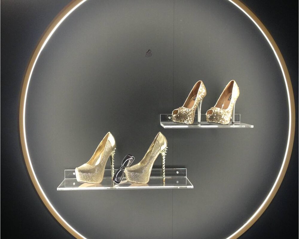 Ringleuchte GIRATA VERTICAL setzt Schuhe in Szene | © SATTLER Innenkreis der Ringleuchte GIRATA als Präsentationsfläche für goldene Schuhe & schwarze Sonnenbrille auf Regalen vor einer schwarzen Wand. | © SATTLER