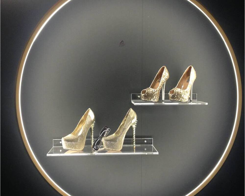 Ringleuchte GIRATA VERTICAL setzt Schuhe in Szene | © SATTLER Innenkreis der Ringleuchte GIRATA als Präsentationsfläche für goldene Schuhe & schwarze Sonnenbrille auf Regalen vor einer schwarzen Wand. | © SATTLER