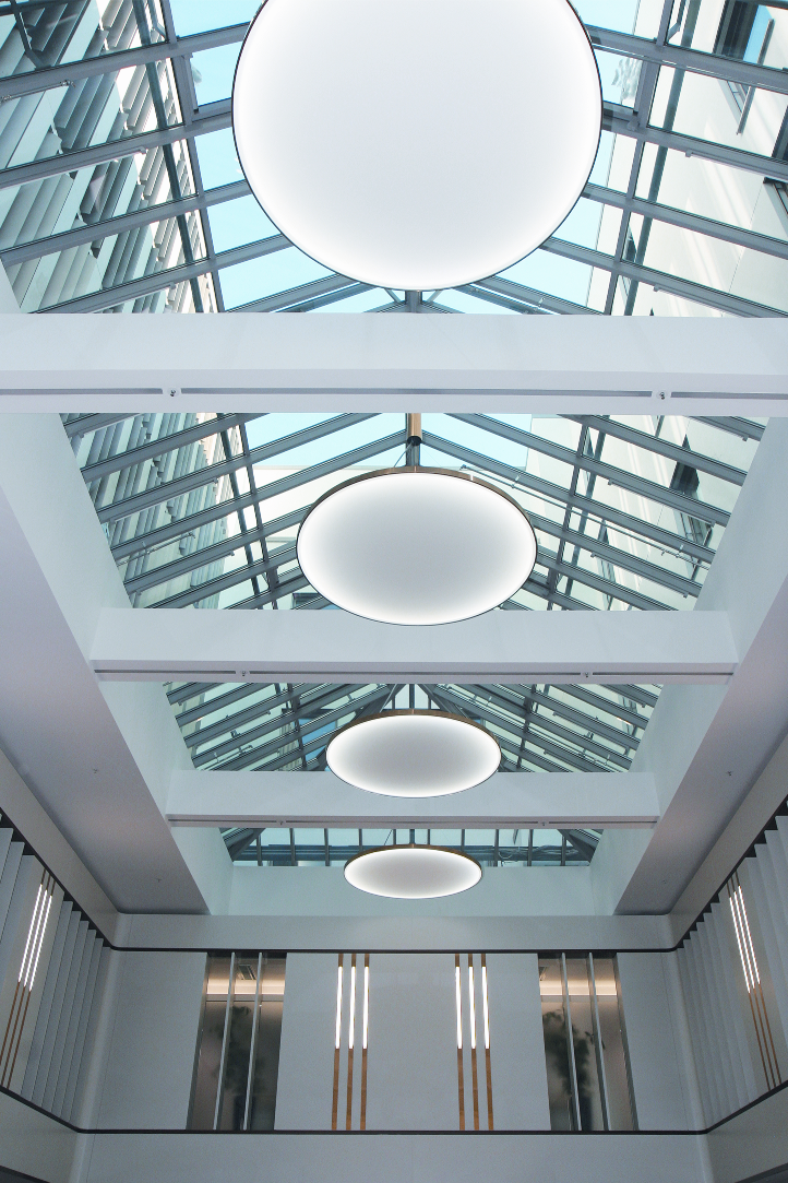 © Jens Laser, Hell und Dunkel Blick von unten auf runde flächige Leuchten, abgependelt in einem Atrium mit spitzem Glasdach. | © Jens Laser, Hell und Dunkel
