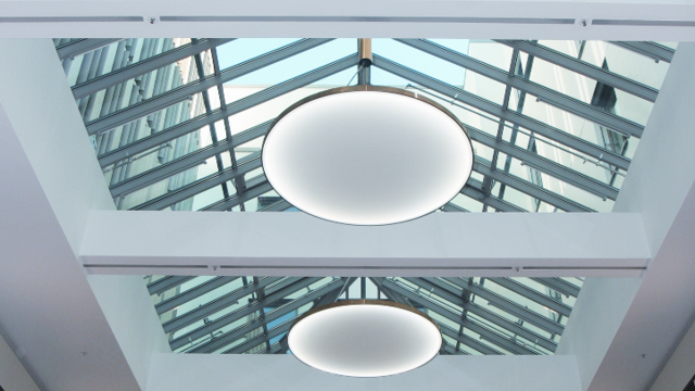 View from below of round, flat luminaires suspended in an atrium with a pointed glass roof. | © SATTLER