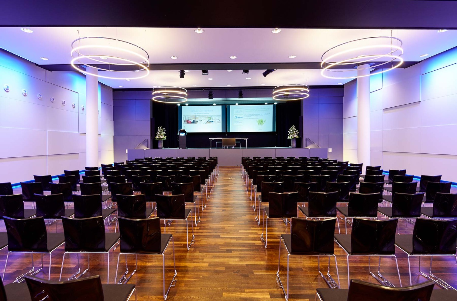 Bananacity event room in Switzerland with a view of the stage, block seating in front of it. Room with white walls coloured purple by light, an INSIEME ring chandelier hangs in all corners of the room.