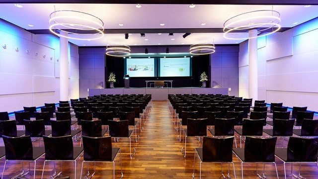 Bananacity event room in Switzerland with a view of the stage, block seating in front of it. Room with white walls coloured purple by light, an INSIEME ring chandelier hangs in all corners of the room. | © http://www.bananacity.ch/restaurant/festsaal/