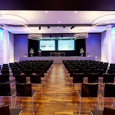 Bananacity event room in Switzerland with a view of the stage, block seating in front of it. Room with white walls coloured purple by light, an INSIEME ring chandelier hangs in all corners of the room.