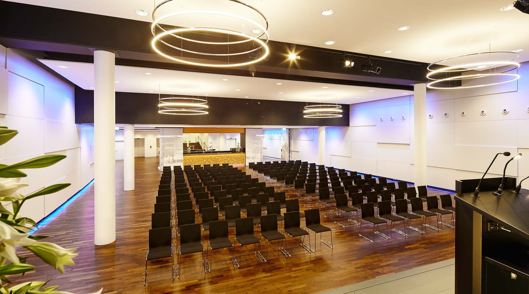 Bananacity Eventsaal mit 4 INSIEME Ring-Kronleuchter | © http://www.bananacity.ch/restaurant/festsaal/ Veranstaltungsraum der Bananacity in der Schweiz mit Blick von der Bühne auf den Saal mit Blockbestuhlung und runden weißen Säulen. Raum hat weiße Wände, die durch Licht lila eingefärbt sind, in allen 4 Raumecken hängt ein INSIEME Ring-Kronleuchter. | © http://www.bananacity.ch/restaurant/festsaal/