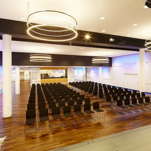 Bananacity event room in Switzerland with a view from the stage onto the hall with block seating and round white pillars. The room has white walls coloured purple by light and an INSIEME ring chandelier hangs in all 4 corners of the room.