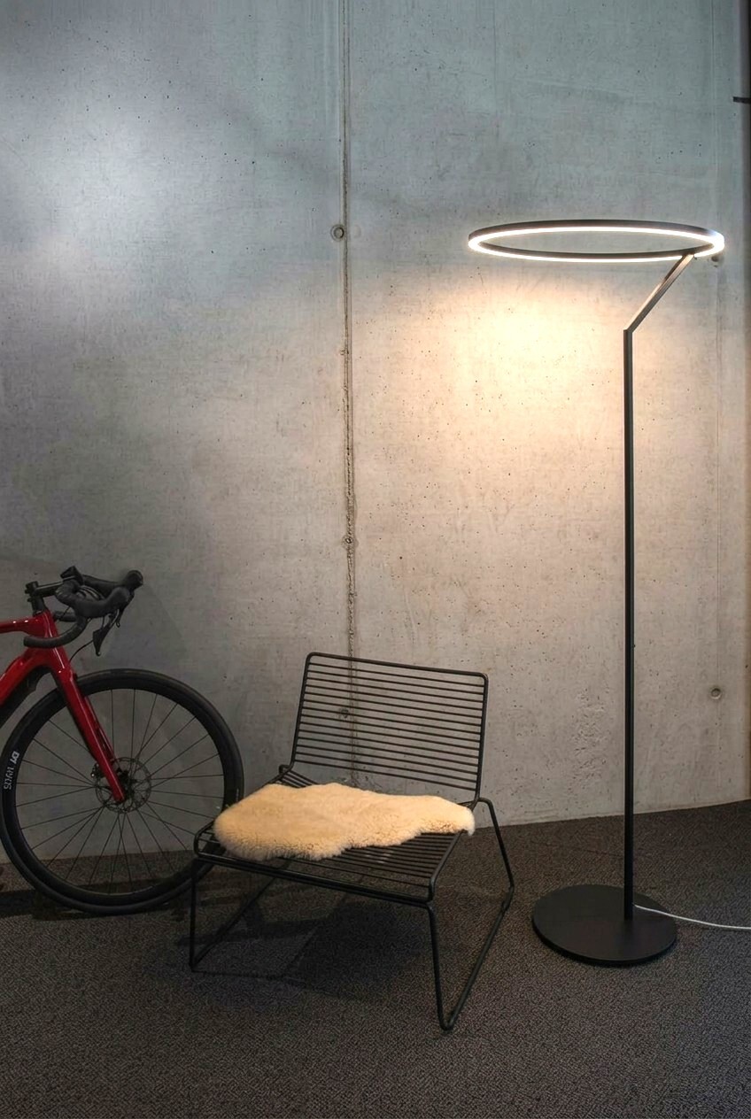 A concrete wall with a red bicycle standing to the left of it, next to it a modern metal chair, and next to that a large floor lamp, the Insospeso Floor: a straight, slender metal base tapers upwards into a luminous ring that casts light downwards. 