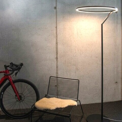 Betonwand an der von links ein rotes Fahrrad steht, daneben ein moderner Metallstuhl, daneben steht eine große Stehleuchte, die Insospeso Floor: Gerader schmaler Metallfuß mündet oben in einen leuchtenden Ring, der nach unten strahlt. 
