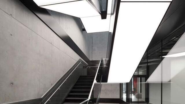 Modern staircase in an exposed concrete building, integrated luminous ceilings run along the underside of the staircase in square, divided strips and in turn illuminate the steps and corridors below. | © Brigida González