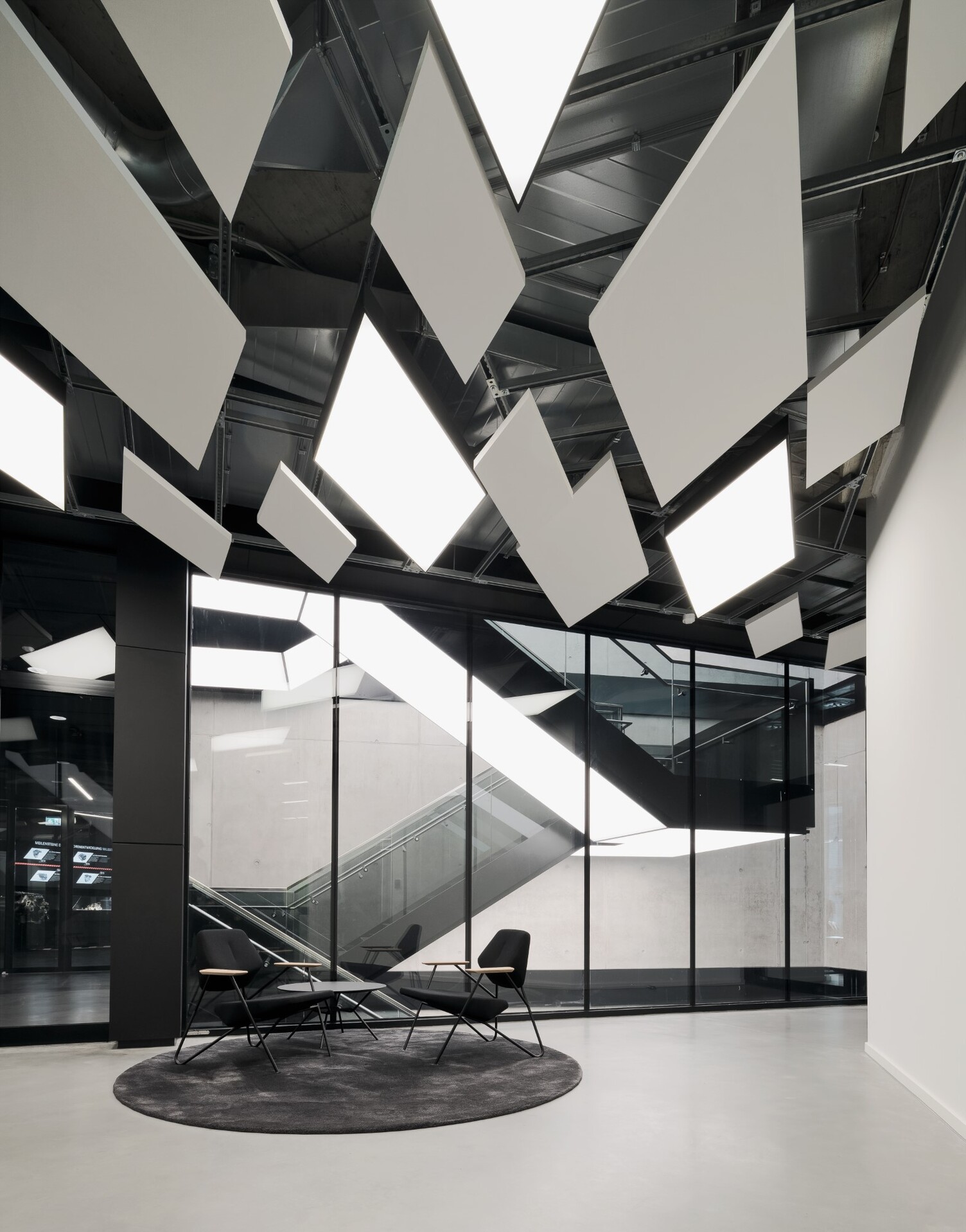 View from a modern waiting area with two chairs and acoustic elements on the ceiling onto a modern stairwell in an exposed concrete building. Integrated illuminated ceilings from SATTLER run along the underside of the stairs in square, divided strips and in turn illuminate the steps and corridors below.