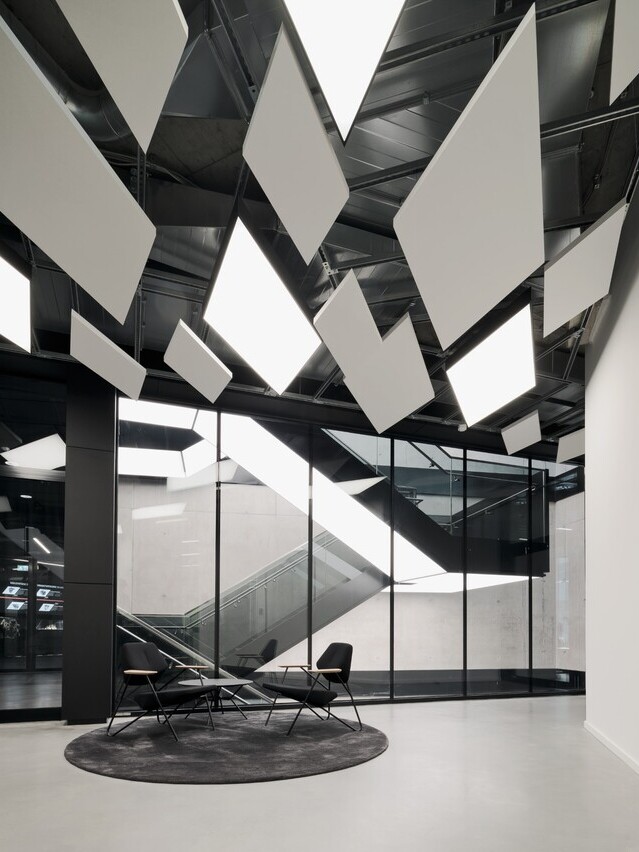 Blick von einem modernen Wartebereich mit zwei Stühlen und Akustikelementen an der Decke auf ein modernes Treppenhaus in einem Sichtbetongebäude, auf der Treppenunterseite verlaufen integrierte Lichtdecken von SATTLER in viereckigen unterteilten Bahnen und beleuchten wiederum die jeweils darunter liegenden Treppenstufen und Flure. | © Brigida González