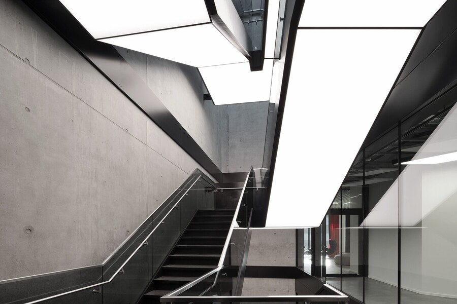 Modern staircase in an exposed concrete building, integrated luminous ceilings run along the underside of the staircase in square, divided strips and in turn illuminate the steps and corridors below.