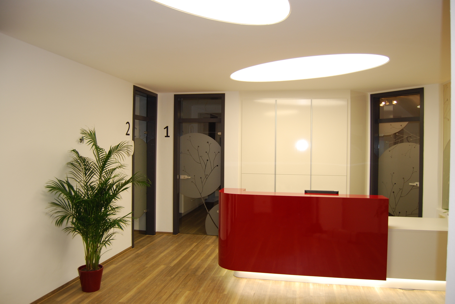 Rote Hochglanz-Empfangstheke über der eine ovale Einbaulichtdecke von SATTLER die Theke beleuchtet, daneben sind Glastüren abgebildet mit Wandbeschriftung für Raumbezeichnung 1 und 2.