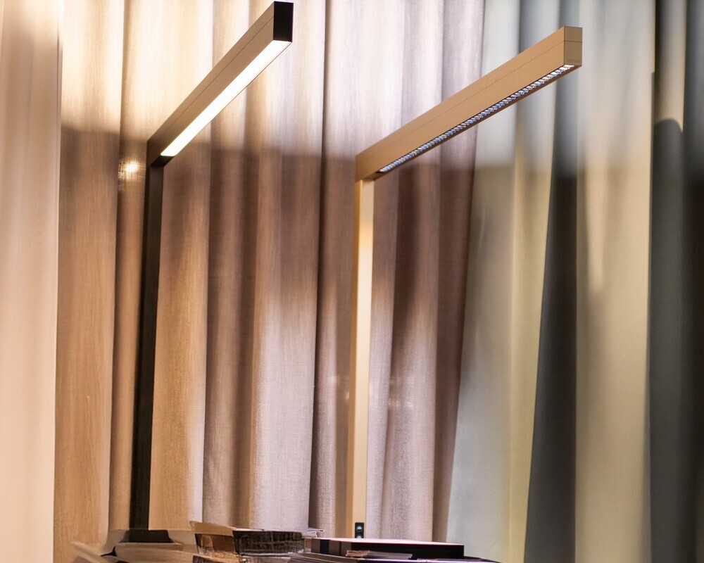 Zwei Diffusoren zur Auswahl: PRISM oder LENS Zwei lineare Stehleuchten der Serie PALADIO FLOOR, links in schwarz mit Optik-Version PRISM, rechts in Champanger Gold mit der Optik-Version LENS, stehen an der kurzen hinteren Seite eines rechteckigen schwarzen Stehtisches vor einer Vorhangwand.