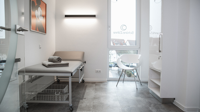 Praxisbehandlungsraum der Praxis Dr. Hensel mit Liege hinter der an der Wand eine lineare Wandleuchte PALADIO WALL angebracht ist, die direkt und indirekt leuchtet. | © Studio Troncone