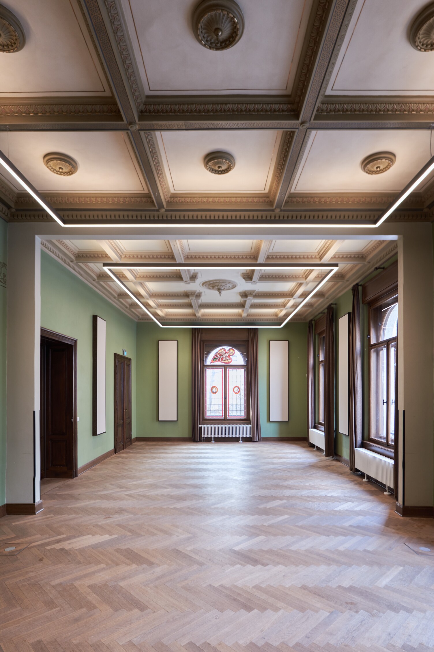 The historic room of the St Thomas Choir (Villa Thomana Chamber Music Hall, Leipzig) features herringbone parquet flooring, ornate antique stained-glass windows, green walls and stucco elements on the ceilings. Suspended from the ceiling is a large square frame light, the PALADIO PRISM system light, which complements the shape of the room and the squares on the ceiling. | © W&V-Architekten