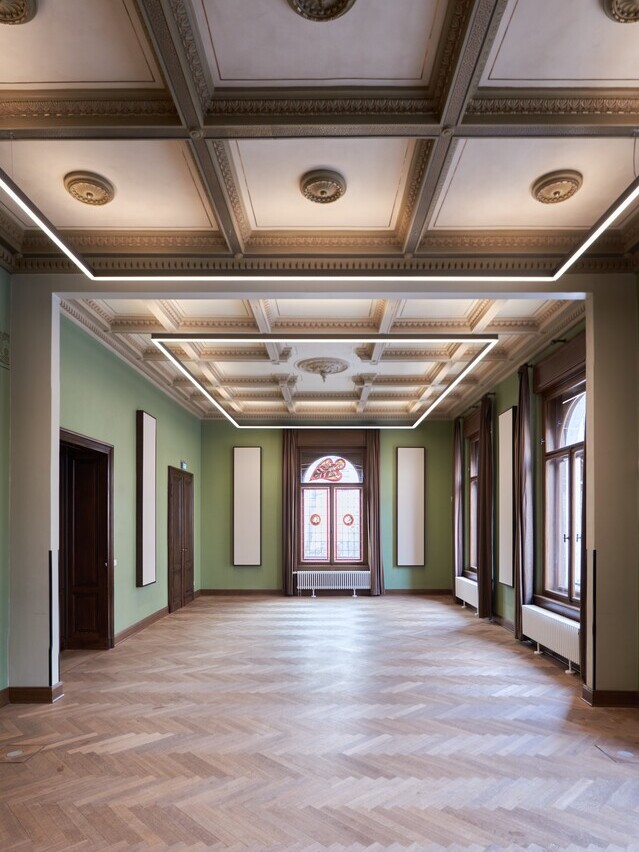 Historischer Raum des Thomanerchor (Kammermusiksaal Villa Thomana, Leipzig) mit Fischgrät-Parkett, verzierte alte Glasfenster, grünen Wänden und Stuckelementen an den Decken. Entlang der Decke ist eine große viereckige Rahmenleuchte, PALADIO PRISM Systemleuchte, abgependelt, die der Form des Raumes und den Quadraten an der Decke schmeichelt. | © W&V-Architekten