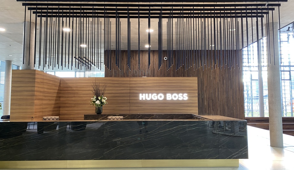 Dunkler eckiger edler Marmorempfangstresen mit Holzrückwand auf der leuchtend weiß das Hugo Boss Logo steht. Über dem Empfangsbereich sind parallel mehrere PALADIO TRACK Stromschienen an der Decke angebracht, von denen lange zylindrische Leuzchten abgependelt sind.