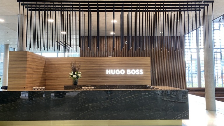 Dark, angular, elegant marble reception desk with a wooden back panel bearing the Hugo Boss logo in bright white. Above the reception area, several PALADIO TRACK tracks are mounted in parallel on the ceiling, from which long cylindrical lights are suspended.