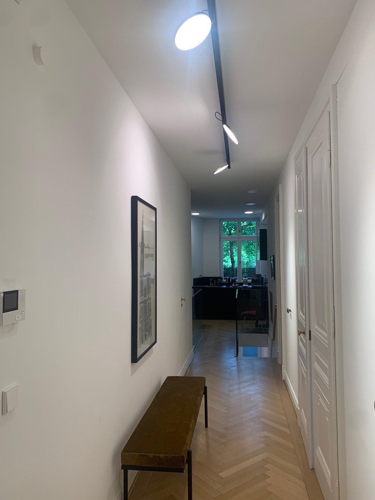 Hallway with parquet flooring leading to an office. The hallway is illuminated by a linear PALADIO TRACK surface-mounted luminaire in black, which houses several round, flat AVVENI SPOT heads that are aligned in different directions and illuminate the hallway.
