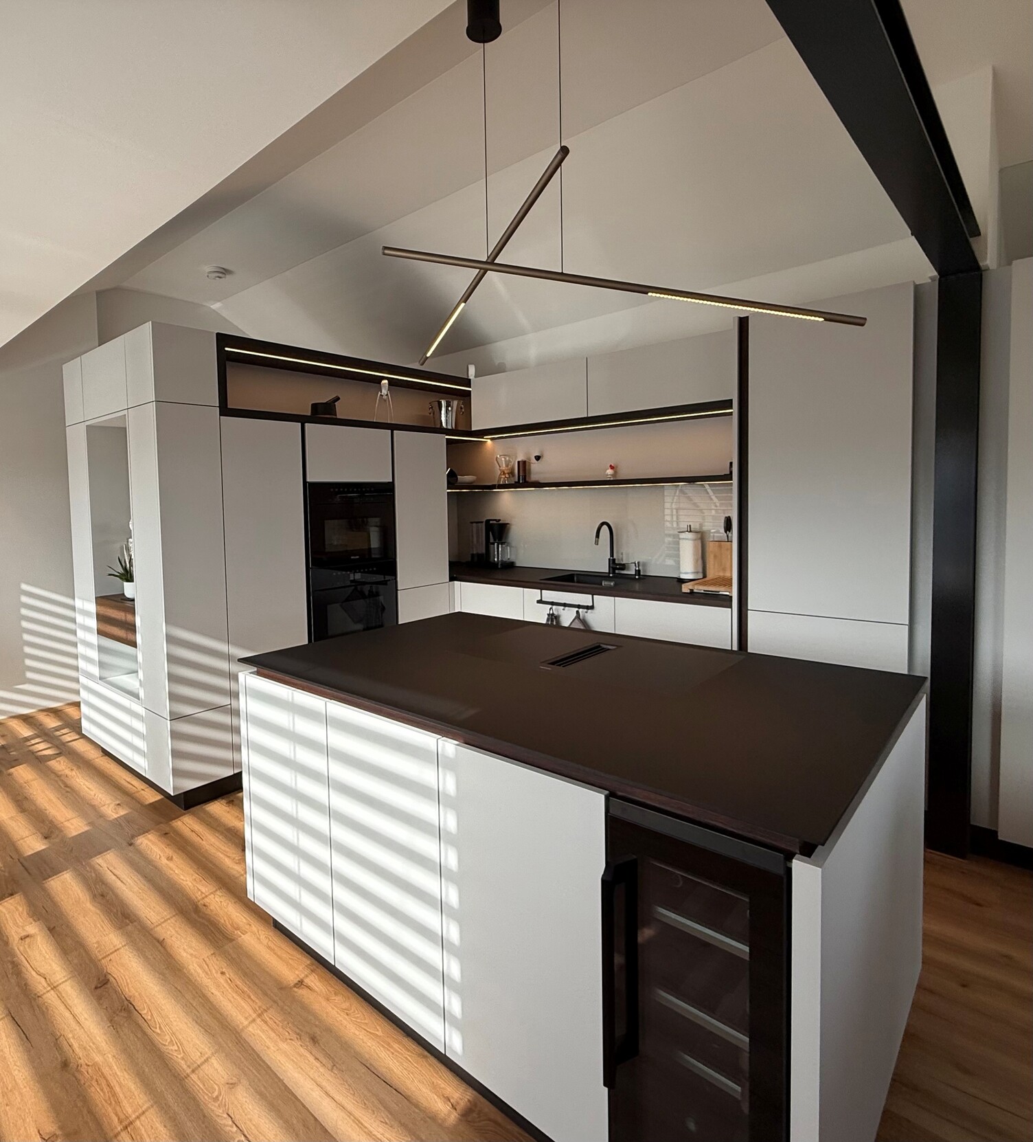 Kitchen lamps. Modern private open kitchen with wooden flooring, modern furniture, kitchen cabinets and kitchen island in the centre are white with black elements. Above the freestanding kitchen island are two cylindrical, horizontal PALITO pendant lights.