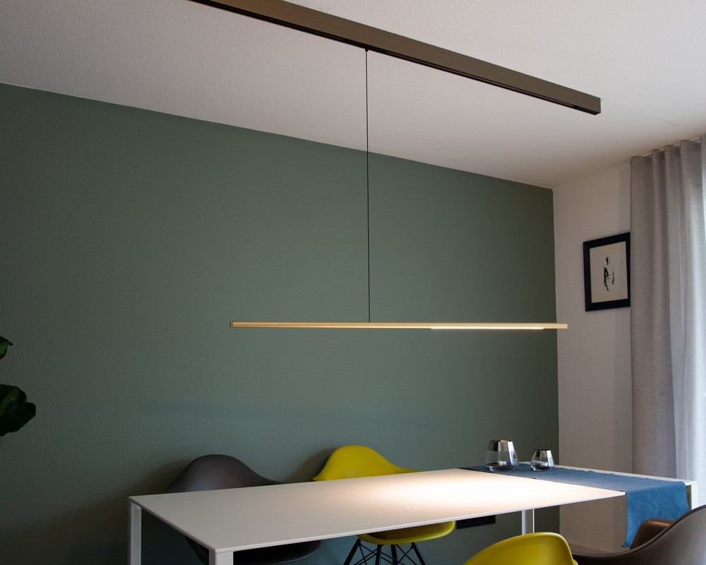 PALITO für PALADIO TRACK (Stromschienensystem) | © Horst Mohring Esszimmer mit weißem rechteckigen Tisch um den gelbe und graue Schalenstühle stehen, dieser steht vor einer grünen Wand. Auf dem Tisch ist ein Lichtkegel zu sehen, der von einer zylindrischen horizontal hängenden Leuchte abstrahlt, diese ist wiederum von einer Aufbaustromschiene von der Decke abgependelt. | © Horst Mohring