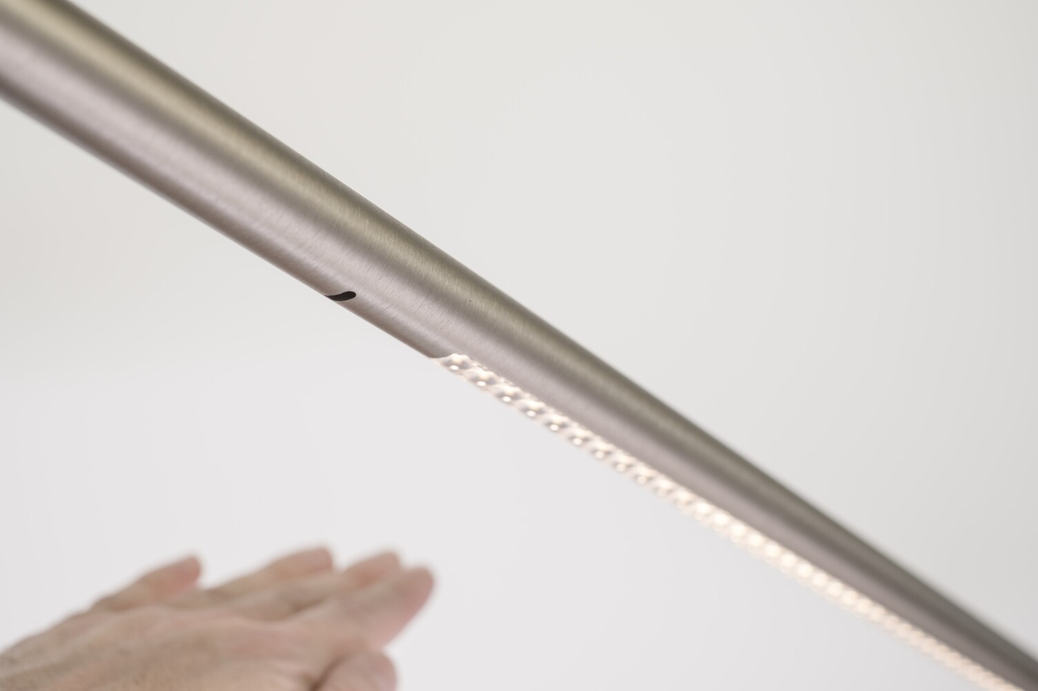 Zylindrische schräg horizontal verlaufende Aluminiumleuchte, unter einem kleinen Rohrloch ist eine Hand zu sehen, im weiteren Verlauf der zylindrischen Leuchte sind im Bild rechts leuchtende Linsen zu sehen. Bild stellt im gesamten die Gestensteuerung wie Dimmung der Leuchte dar.