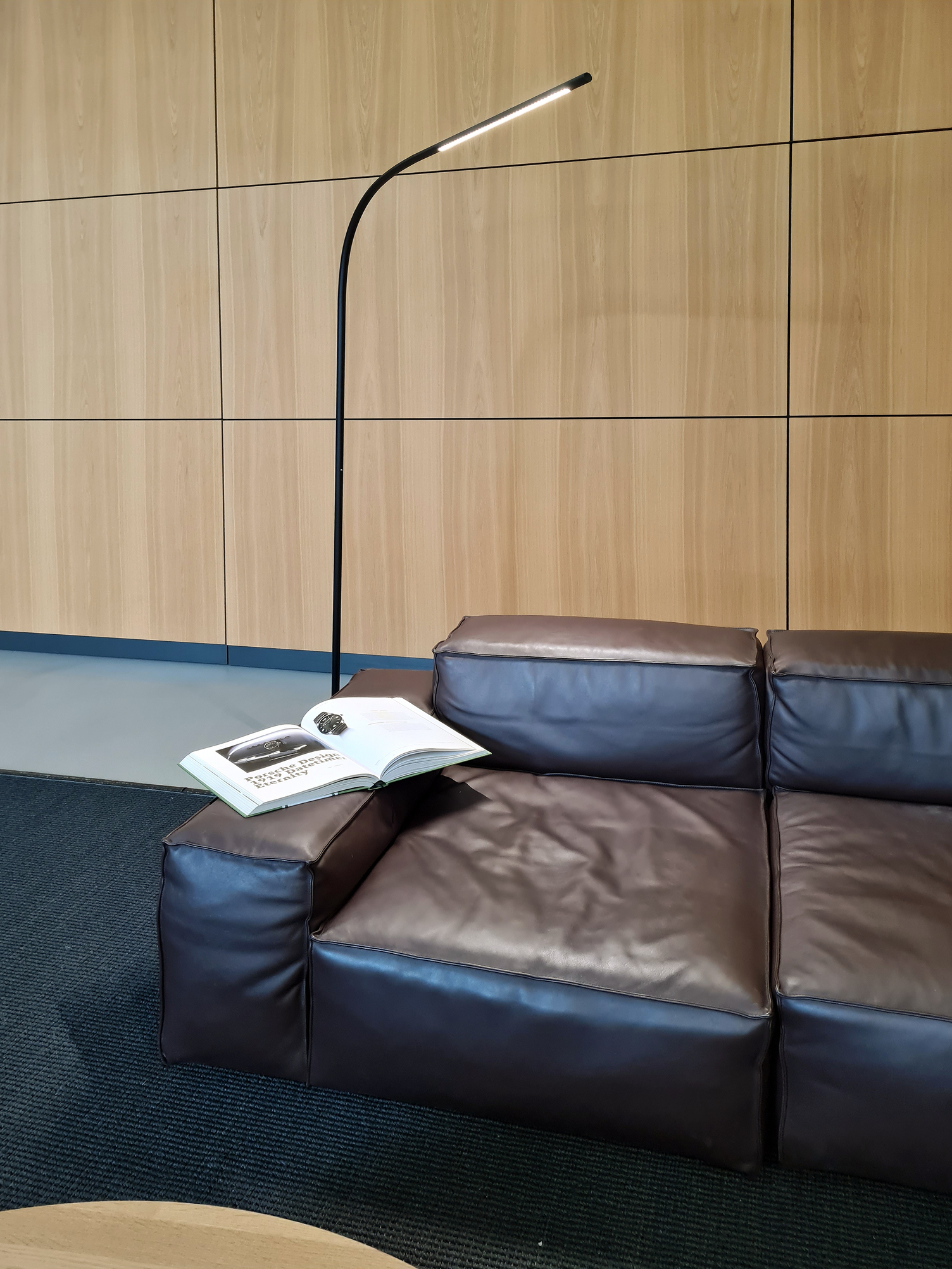 Stehleuchte PALITO FLOOR schwarz (gebürstet, eloxiert) Braunes Ledersofa vor einer Holzwand, links auf dem Sofarm liegt ein aufgeschlagenes Buch, dass von der gebogenen Stabstehleuchte beleuchtet wird.