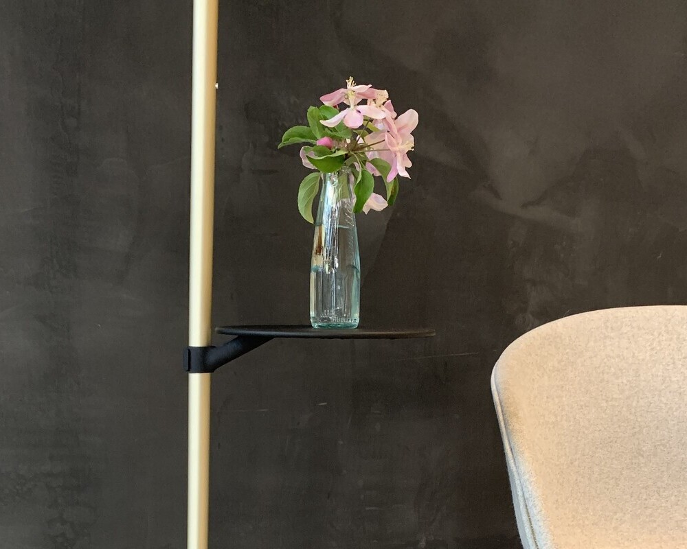 Optionaler Ablagetisch Ausschnitt der Stehleuchte PALITO FLOOR links im Bild, zeigt sie in Champagner Gold mit schwarzem runden Ablagetischchen auf dem eine Glasvase mit rosa Blumen steht. Rechts daneben ist eine weiße Stuhllehne zu sehen. Hintergrund: schwarze Wand.