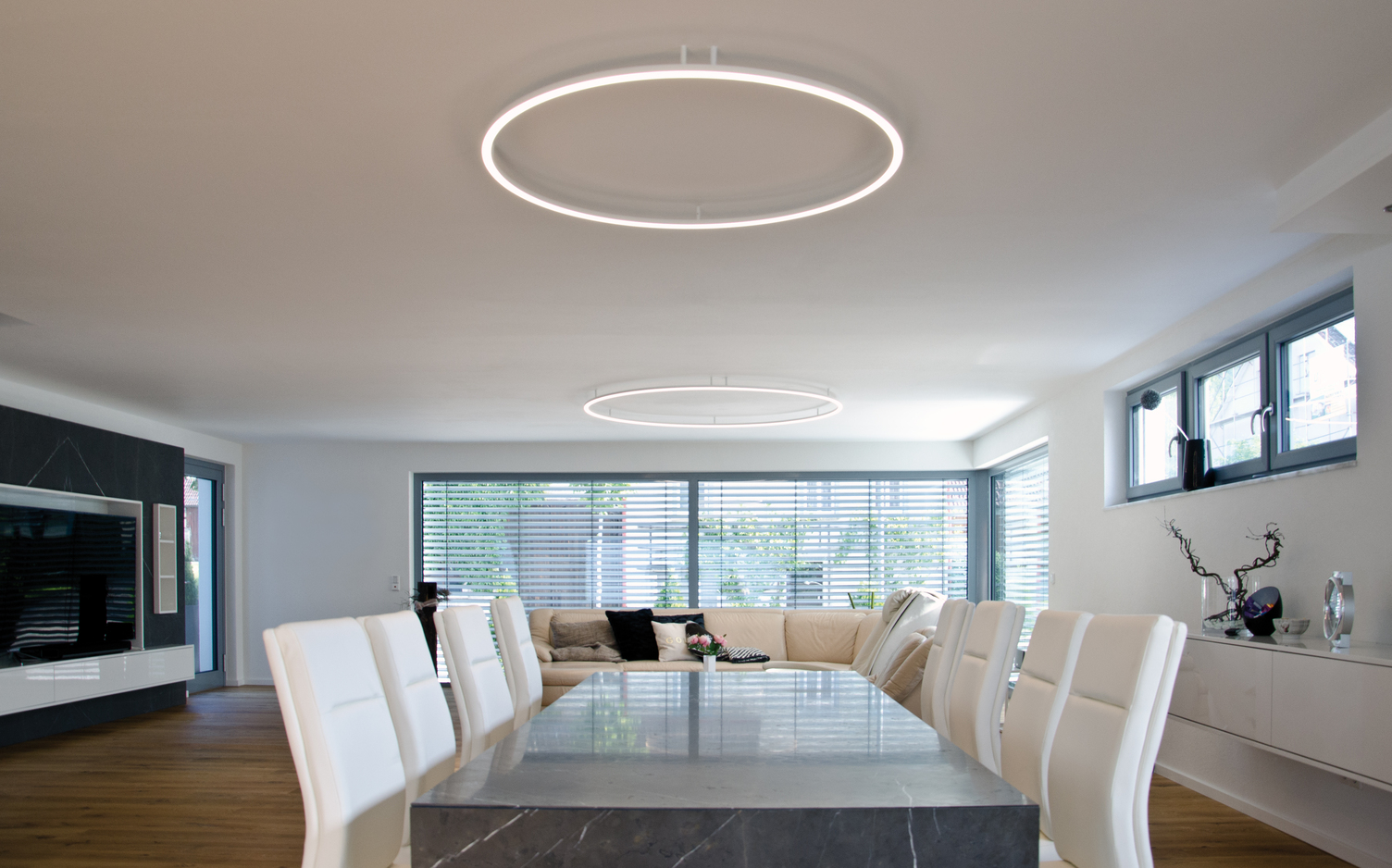 Modernes Wohn- und Esszimmer mit schwarzem Marmortisch, weißen Lederstühlen, sowie weißem Ledersofa, über jeweiligen Bereichen Anbau-Ringleuchte (ROTOLO).