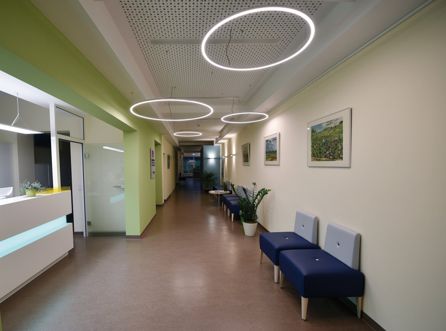 Krankenhaus- oder Praxisflur mit vereinzelten Sitzgelegenheiten und mehreren verteilten von Decke abgehangenen Ringleuchten.