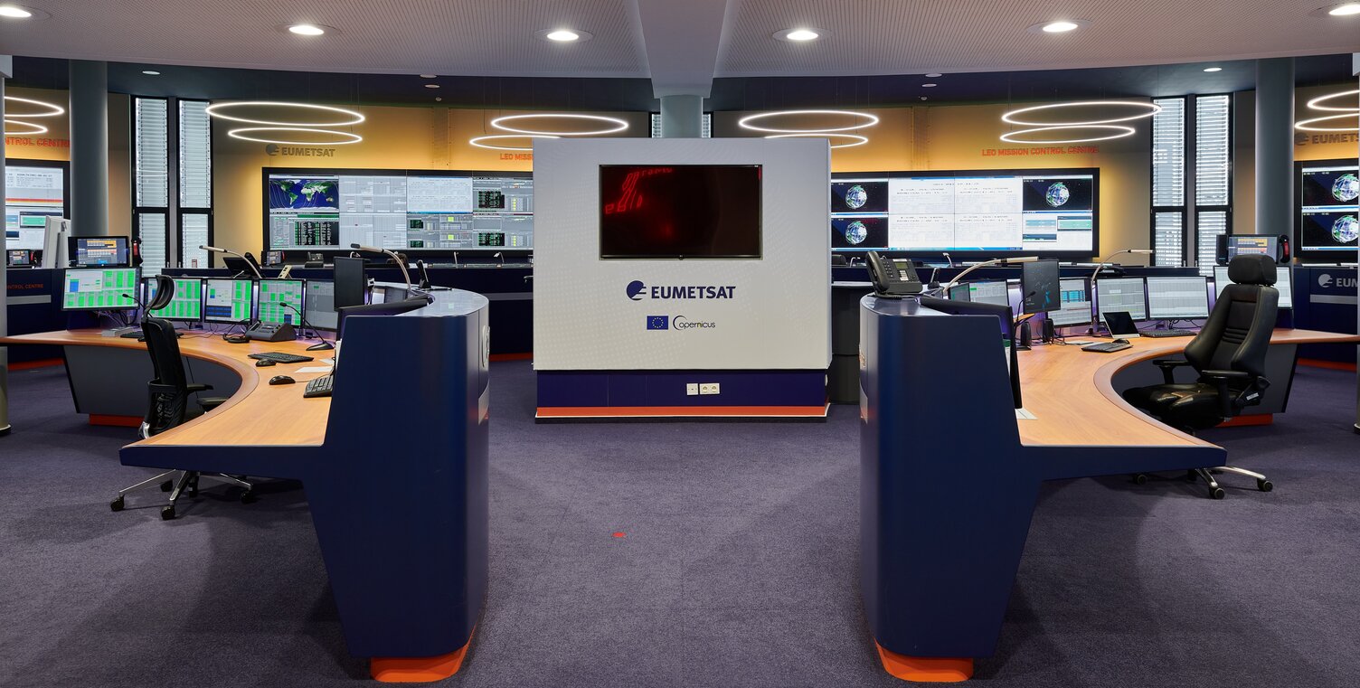 Ein Gang führt in ein Kontrollzentrum mit mehreren Tischen und großen Bildschirmen. Bildschirmnah sind mehrere Ringleuchten von der Decke abgehangen. | © EUMETSAT