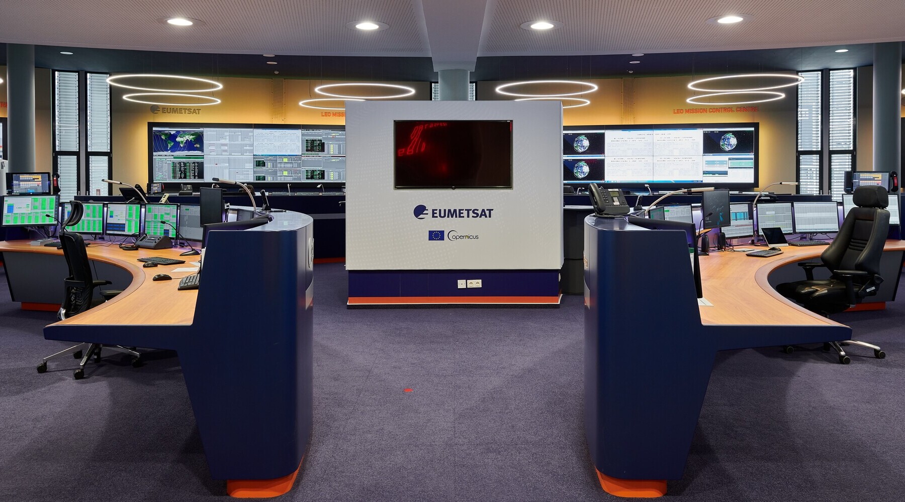 © EUMETSAT Ein Gang führt in ein Kontrollzentrum mit mehreren Tischen und großen Bildschirmen. Bildschirmnah sind mehrere Ringleuchten von der Decke abgehangen. | © EUMETSAT