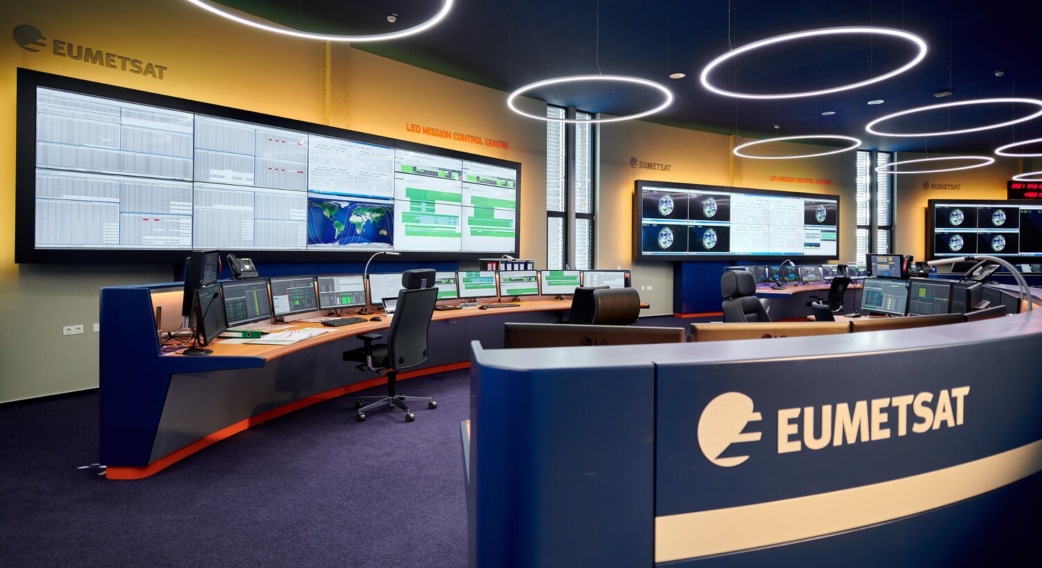 © EUMETSAT Kontrollzentrale von EUMETSAT mit mehreren großen Bildschirmen in einem gelb, blau gehaltenem Raum mit großen schmalen Ringleuchten von der Decke abgependelt. | © EUMETSAT