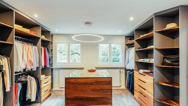Modern dressing room with shelves and wardrobes with clothes to the left and right of the room and a wooden table block in the centre above which hangs a SLIM DIRECT ring light. | © Frank Luger