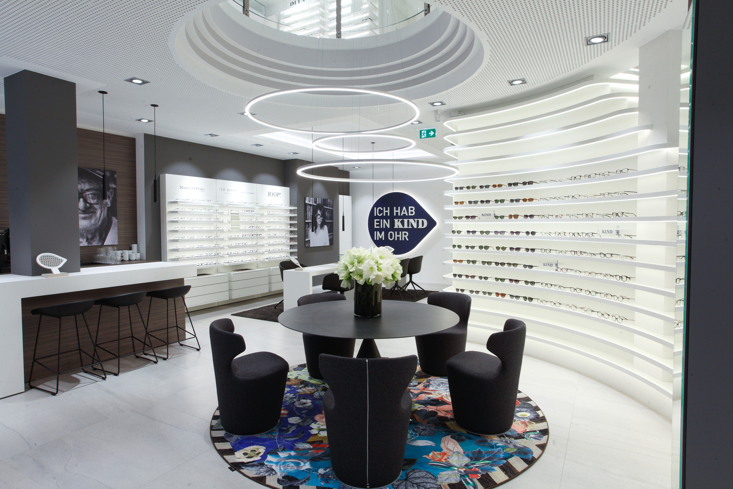 3 SLIM ring light in the centre of the room above a round black consultation table on the walls Displays with various spectacles and an advertising slogan for KIND hearing aids.