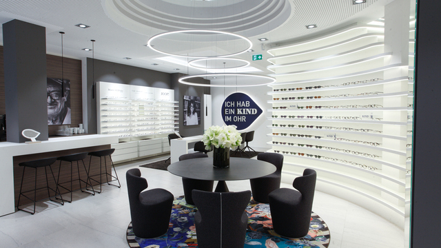 3 SLIM ring light in the centre of the room above a round black consultation table on the walls Displays with various spectacles and an advertising slogan for KIND hearing aids. | © Seydlitz GmbH