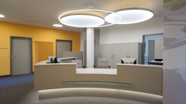 One filigree SLIM DIRECT LED ring light by SATTLER above the reception desk, with two Highmoon Flat luminaires behind, modern lighting design in a medical practice foyer.