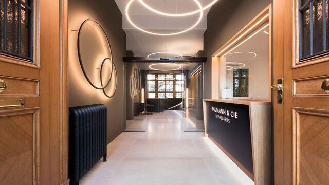 A historic double wooden entrance door with ornaments provides a view of a hallway decorated in wood and earth tones with decoratively arranged Slim Wall opposite the reception area of Baumann Cie bank and Slim direct and Slim indirect ring lights used for basic lighting. | © Andreas Zimmermann Fotografie