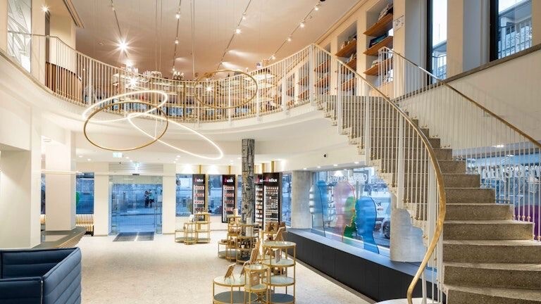 View from the interior of Köln Tourismus to a staircase leading to the first floor and the glass entrance; in the centre of the room, a light object made of SLIM ring lights attracts attention.
