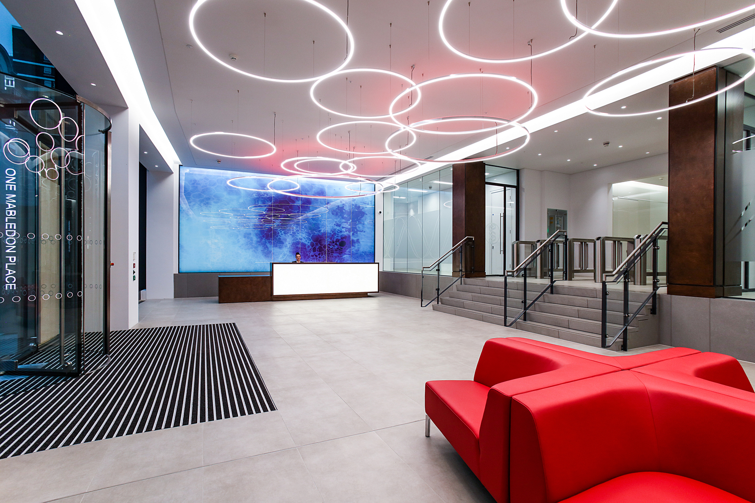 The entrance area of One Mabledon Place in London shows a tiled entrance area above which several SLIM ring lights are suspended. At one end of the room is a reception desk behind which hangs a large blue picture, a red leather sofa on the other side of the room, stairs lead up one level from the glass entrance door.