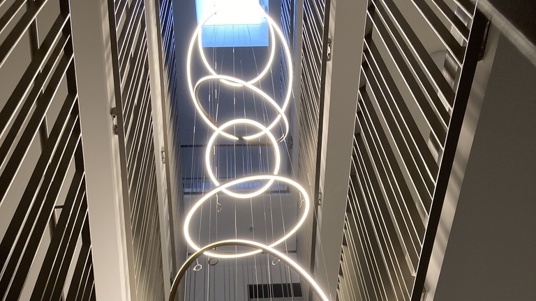 Maßgeschneiderter Kronleuchter fürs Treppenhaus: Blick von einem Treppenauge unten in eine Ansammlung von 7 schmalen Ringleuchten SLIM, die sich im Treppenauge nach oben schlängeln. | © SATTLER