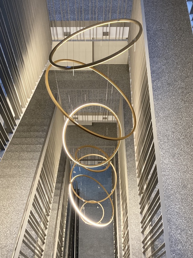 Blick nach unten auf eine Sonderleuchte, wie ein moderner Kronleuchter bestehend aus mehreren direkt leuchtenden Ringleuchten, in einem Treppenauge eines Treppenhauses mit mehrläufigen geraden Treppen. | © Via Modular Licht + Gestaltung GmbH