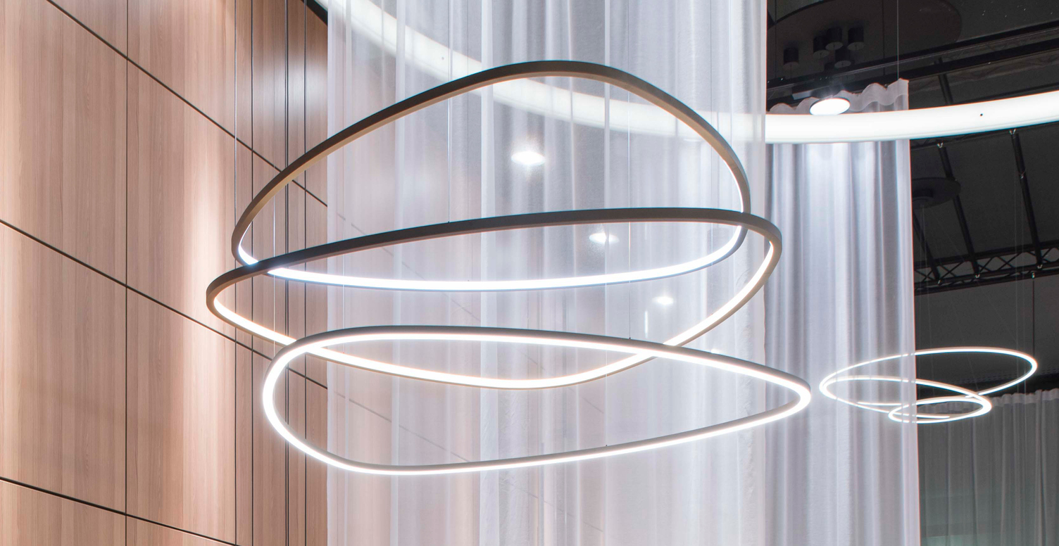 In the foreground, three narrow ring lights in the shape of a rounded triangle are suspended a short distance above each other, with the lowest one shining directly downwards and the two upper ones shining into the centre of the lights. In the background, a brown wooden wall and a slightly transparent white curtain, and a modern TOCCATA ring chandelier.