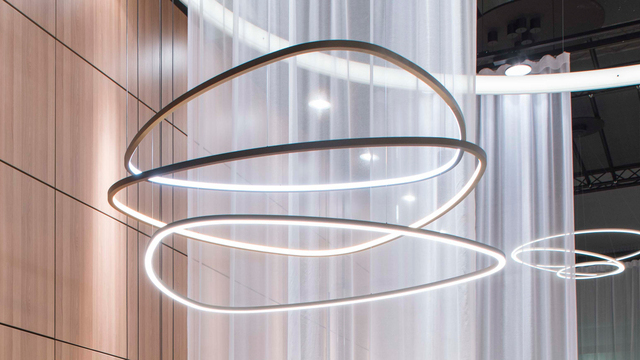 In the foreground, three narrow ring lights in the shape of a rounded triangle are suspended a short distance above each other, with the lowest one shining directly downwards and the two upper ones shining into the centre of the lights. In the background, a brown wooden wall and a slightly transparent white curtain, and a modern TOCCATA ring chandelier. | © SATTLER